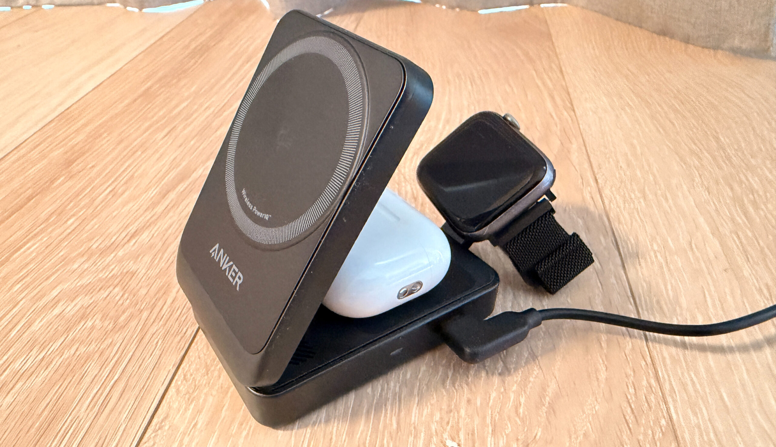 木製テーブルの上に黒い無線充電器、大きな文字で"ANKER"と書かれ、白いイヤホンケースとスマートウォッチが配置されている。