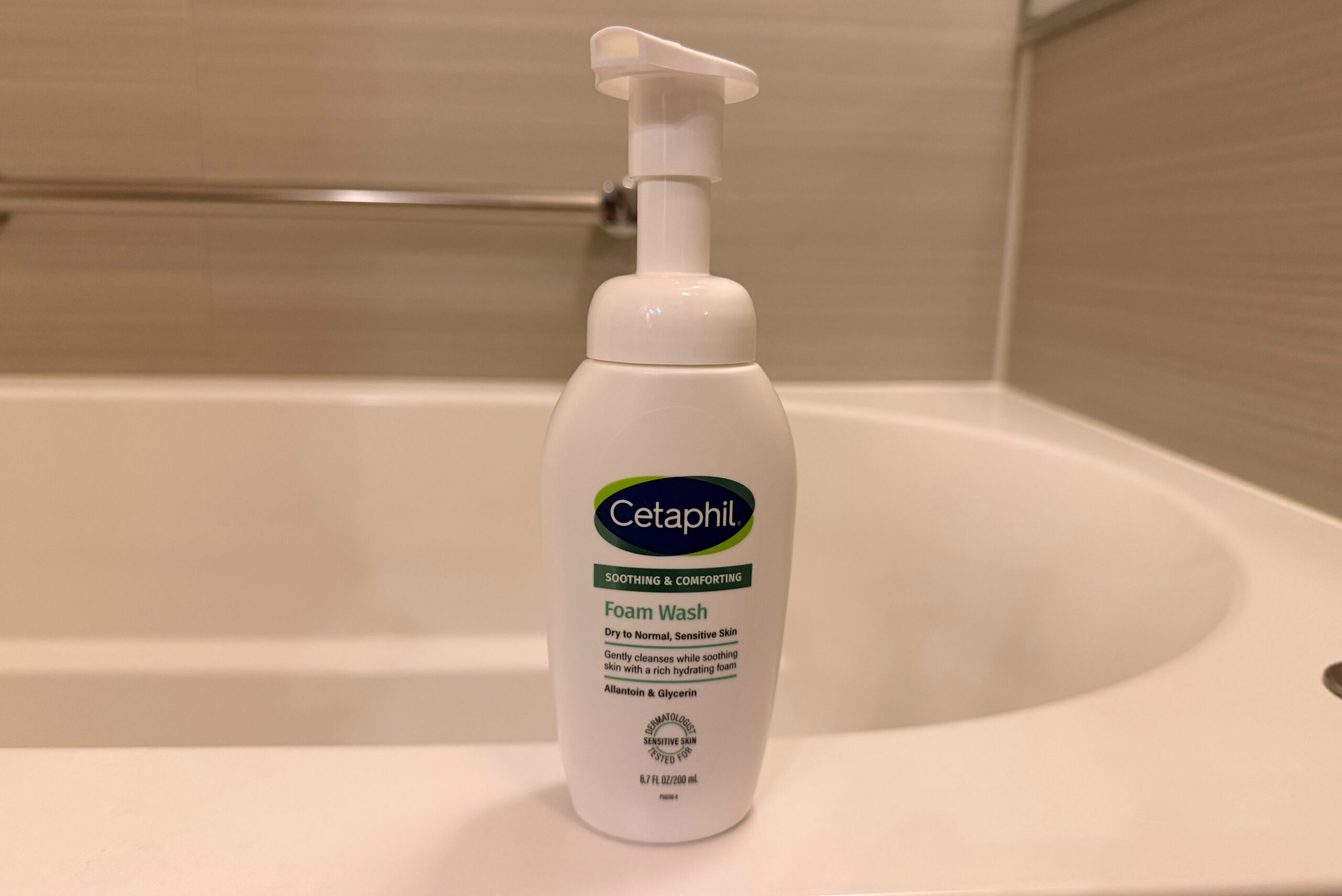 白い「Cetaphil」フォームウォッシュ、浴槽の縁に置かれ、落ち着いたバスルームの背景。ラベルに製品名と説明文が記載。